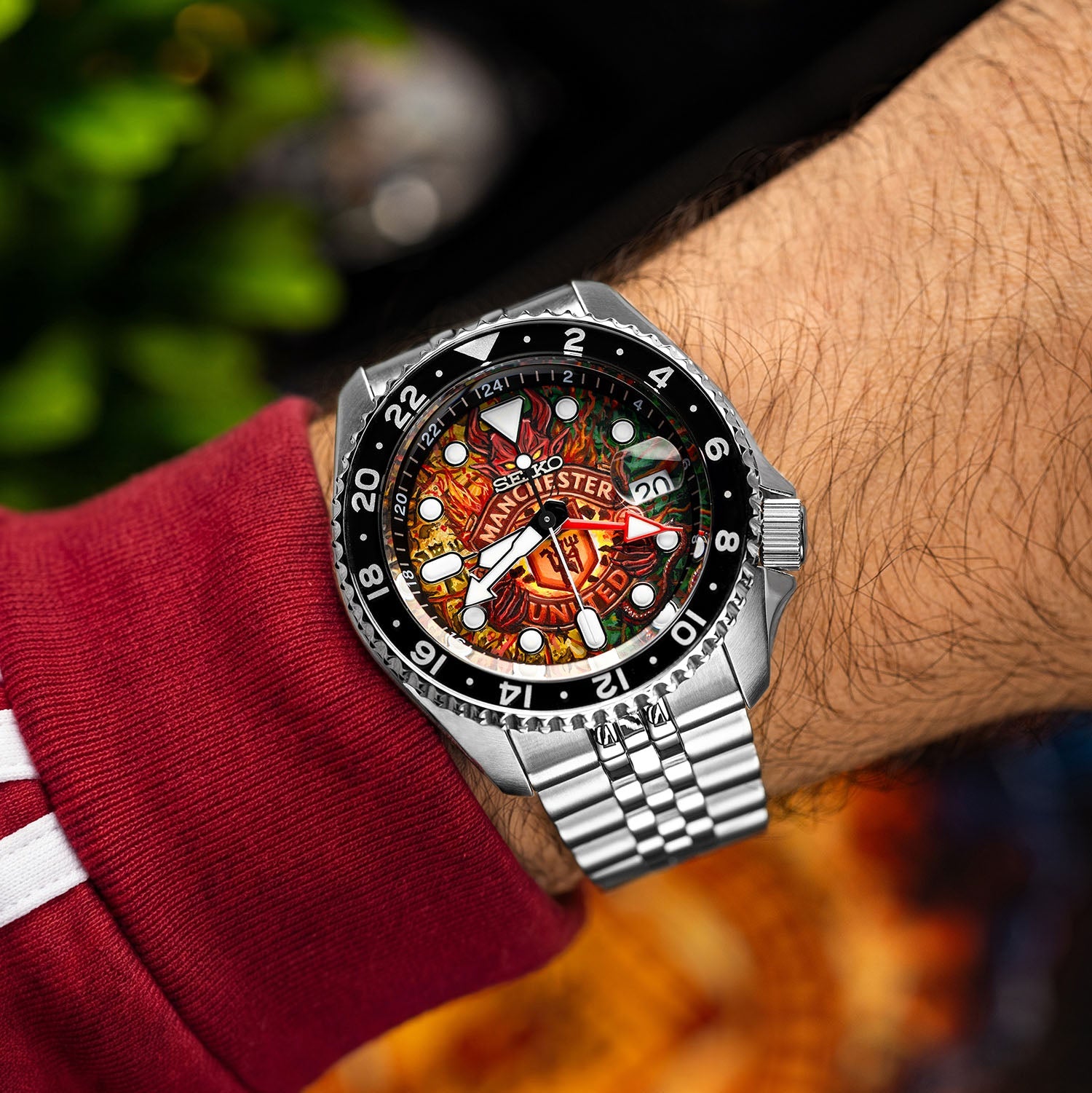 The Red Devils Concept on wrist — hand-painted football crest dial with red and golden flames visible against a red jersey sleeve, 42.5mm stainless steel GMT automatic with black bezel and jubilee bracelet, a bespoke timepiece by IFL Watches.
